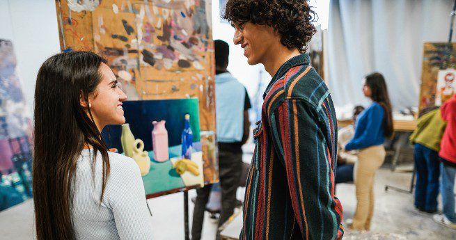 The image shows two young people engaged in conversation while standing in an art studio or gallery. They are smiling at each other, creating a warm and friendly atmosphere. The background features several paintings on easels, with other people also observing and interacting with the artwork.