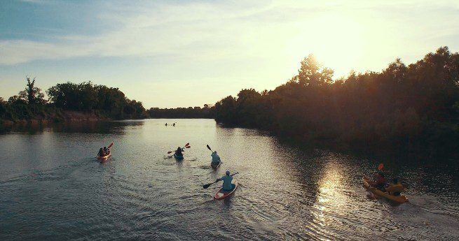 The image shows a group of kayakers paddling down a calm river surrounded by lush greenery. The kayakers are spread out across the water, with the sun setting in the background, casting a warm, golden light over the scene. The riverbanks are lined with trees, and the water reflects the colors of the sunset.