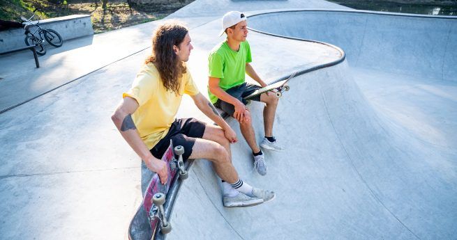 The image shows two young men sitting on the edge of a concrete skatepark, each holding a skateboard. Both are casually dressed, one in a yellow shirt and the other in a green shirt, with shorts and sneakers. One of them is wearing a white cap turned backward.