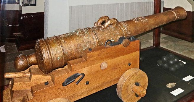 The image shows an old bronze cannon mounted on a wooden carriage with wheels.