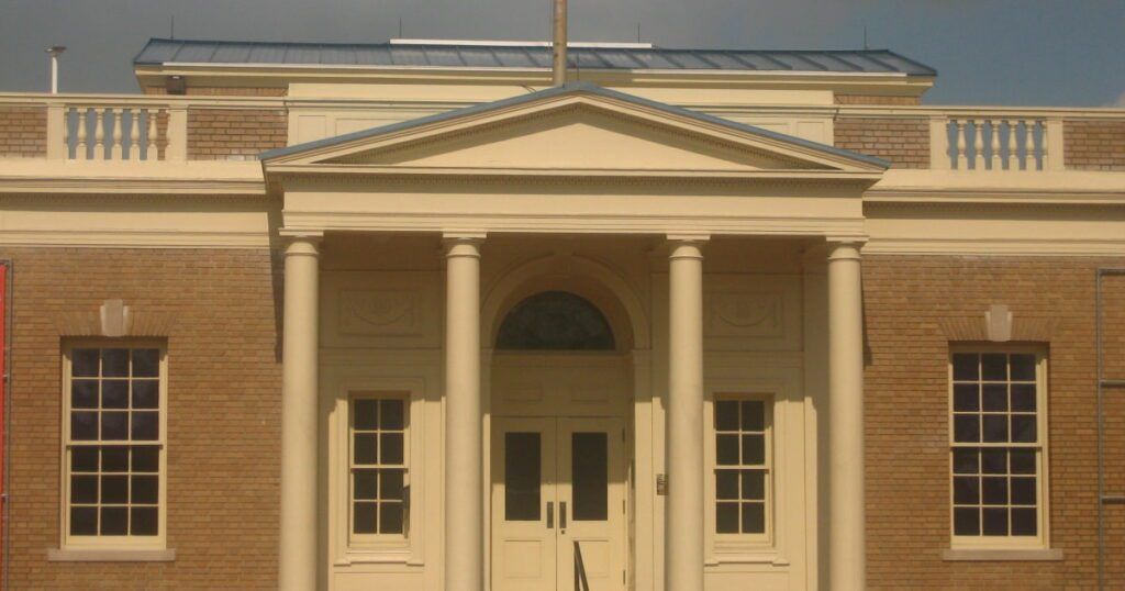 The image shows the front facade of the Matagorda County Museum, designed in a classical architectural style. The building features a symmetrical design with a central entrance framed by four tall columns that support a triangular pediment.