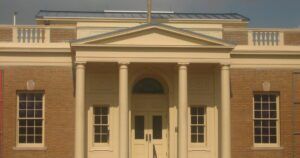 The image shows the front facade of the Matagorda County Museum, designed in a classical architectural style. The building features a symmetrical design with a central entrance framed by four tall columns that support a triangular pediment.