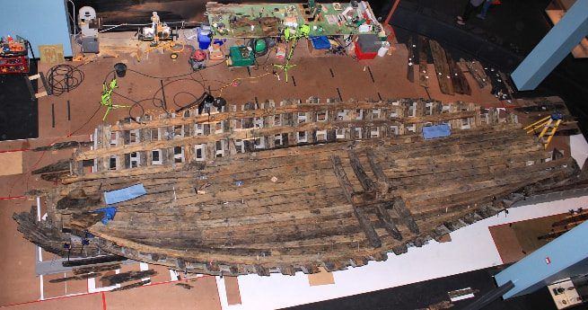 The image shows the well-preserved remains of the La Belle shipwreck, a 17th-century French exploration vessel. The wooden hull is laid out horizontally in a conservation facility, with various tools and equipment surrounding it, indicating ongoing restoration and preservation efforts. The ship's structure, though aged and weathered, remains remarkably intact, showcasing the shipbuilding techniques of the era. The careful arrangement of the site and the focus on detailed conservation work reflect the historical significance of La Belle, which played a crucial role in French exploration in the Americas.