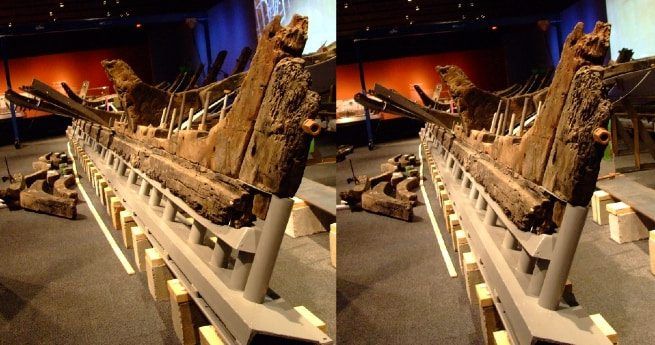 The image displays the side view of the La Belle shipwreck, showcasing the wooden remains of the vessel's hull, which are carefully supported by a metal frame in a museum setting.