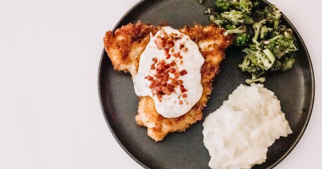 A plate of food featuring a crispy, golden-brown chicken fried steak topped with a generous dollop of creamy white gravy and a sprinkling of bacon bits. The dish is served with a side of fluffy mashed potatoes and a portion of sautéed greens, possibly broccoli, all presented on a dark plate against a white background.