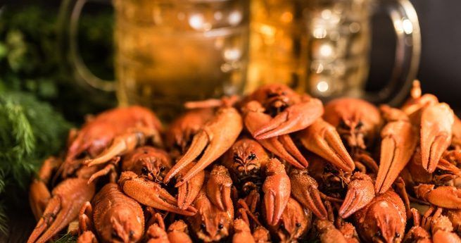 A close-up view of a pile of freshly cooked crawfish, their vibrant red shells glistening under the light. The crawfish are the focal point of the image, with two large, frosty mugs of beer blurred in the background.