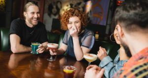 A group of friends is sitting at a bar, enjoying colorful cocktails and laughing together. The scene captures a lighthearted and joyful moment, with a woman in the center playfully covering her mouth while laughing.