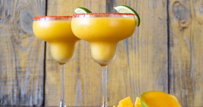 Two frosty mango margaritas are served in classic margarita glasses, each garnished with a slice of lime and a red chili-salt rim. The vibrant yellow-orange color of the drinks contrasts beautifully with the rustic wooden background. Fresh mango slices are placed beside the glasses, emphasizing the tropical flavor of the cocktails.