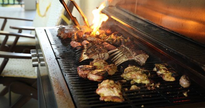 A close-up of a barbecue grill loaded with various meats, including racks of ribs, chicken pieces, and skewers. The grill flames are flaring up, adding a smoky char to the food. The background shows patio furniture, suggesting an outdoor grilling setting.