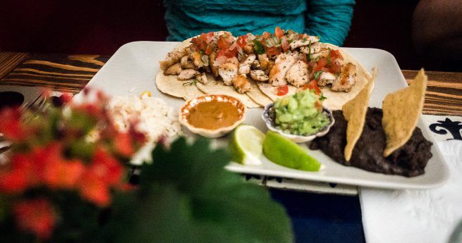 A plate of Mexican food featuring two tacos filled with grilled chicken and topped with pico de gallo. The plate also includes sides of refried black beans garnished with tortilla chips, guacamole, a lime wedge, and a small serving of rice. A small dish of salsa is placed on the plate, and the meal is set on a wooden table with a person in a teal shirt in the background.