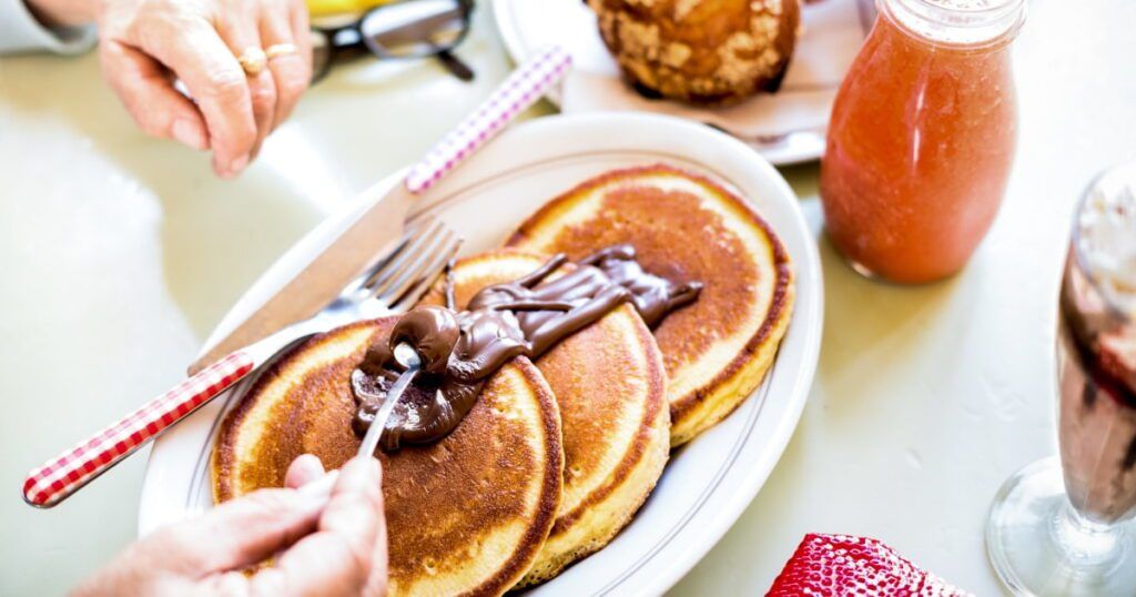 A close-up of a breakfast plate featuring three golden-brown pancakes topped with a generous swirl of chocolate spread.