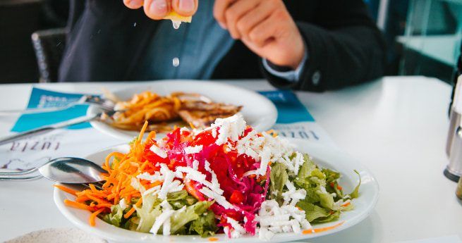 A close-up of a fresh salad topped with shredded cheese, vibrant red cabbage, and grated carrots, served on a white plate. In the background, a person is squeezing a lemon over the salad, with another dish partially visible on the table.