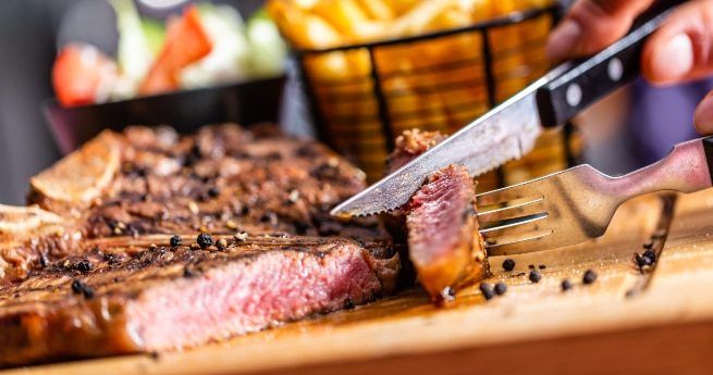 A close-up of a juicy, medium-rare steak being cut with a knife and fork on a wooden board. The steak is seasoned with coarse black pepper, and a basket of golden French fries is visible in the background, along with a bowl of salad.