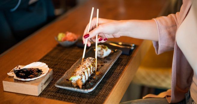 A person with red-painted nails uses chopsticks to pick up a piece of sushi from a rectangular plate. The sushi roll is drizzled with sauce and topped with sesame seeds.