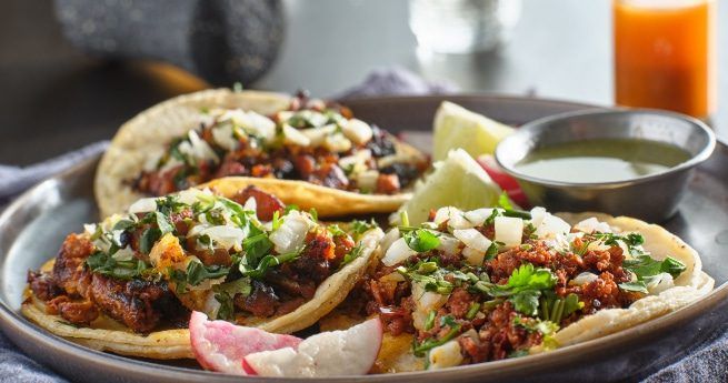 A close-up of a plate with three soft tacos filled with a variety of toppings including seasoned meat, chopped onions, and fresh cilantro. The tacos are garnished with slices of radish and lime wedges. A small bowl of green salsa is placed on the side, with a bottle of hot sauce visible in the blurred background.