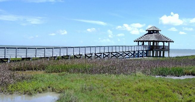 A wooden boardwalk stretches out over a marshy area, leading to a covered observation tower with a pyramid-shaped roof. The structure overlooks a wide expanse of water, with the grassy marshlands in the foreground.