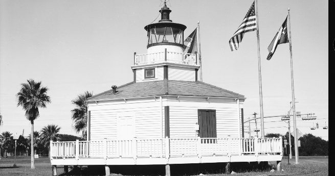 A black-and-white photograph of the Halfmoon Reef Lighthouse, a small, octagonal structure with a cupola housing the light at the top. The lighthouse is made of wood, with a wrap-around deck supported by stilts. The roof is slightly sloped, and the structure has a quaint, historic charm. Three flags are visible to the right of the lighthouse, including the American flag.