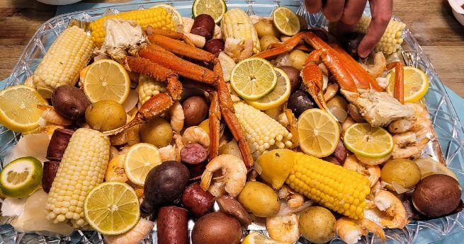 A colorful and appetizing seafood boil spread on a large platter. The dish includes crab legs, shrimp, corn on the cob, small red and yellow potatoes, and sausage, all seasoned and cooked to perfection. Slices of lemon are scattered throughout the platter, adding a fresh, citrusy touch to the meal. A hand is reaching in to grab some of the delicious seafood.