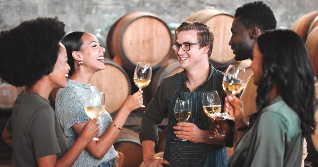 best local wineries and wine bars in port lavaca and victoria-group with wine-5 A group of five people standing together in a winery, smiling and holding glasses of white wine while chatting, with large wine barrels in the background.