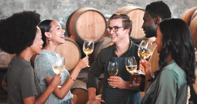 A group of five people standing together in a winery, smiling and holding glasses of white wine while chatting, with large wine barrels in the background.