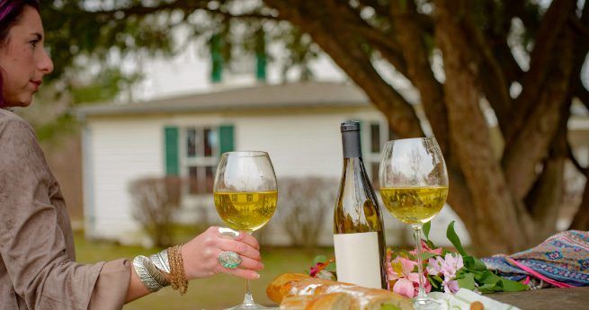 A woman holding a glass of white wine at an outdoor table set with another glass, a bottle of wine, a loaf of bread, and flowers, with a white house and trees in the background.