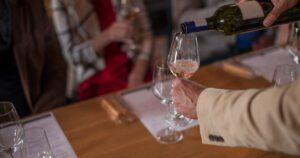 Close-up of a person in a beige jacket pouring rosé wine into a wine glass during a tasting event, with blurred people in the background seated at a table.