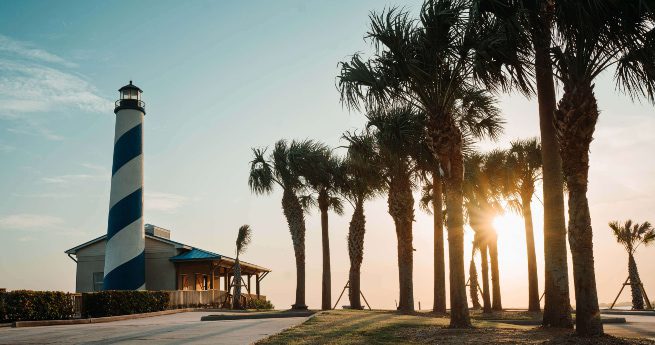 texas vs. florida-beachside view-4 A serene coastal scene featuring a tall lighthouse with blue and white spiral stripes, standing next to a small building with a blue roof. A row of tall palm trees lines the right side of the image, their shadows cast on the ground as the sun sets behind them. The soft sunlight filters through the palms, creating a golden glow.