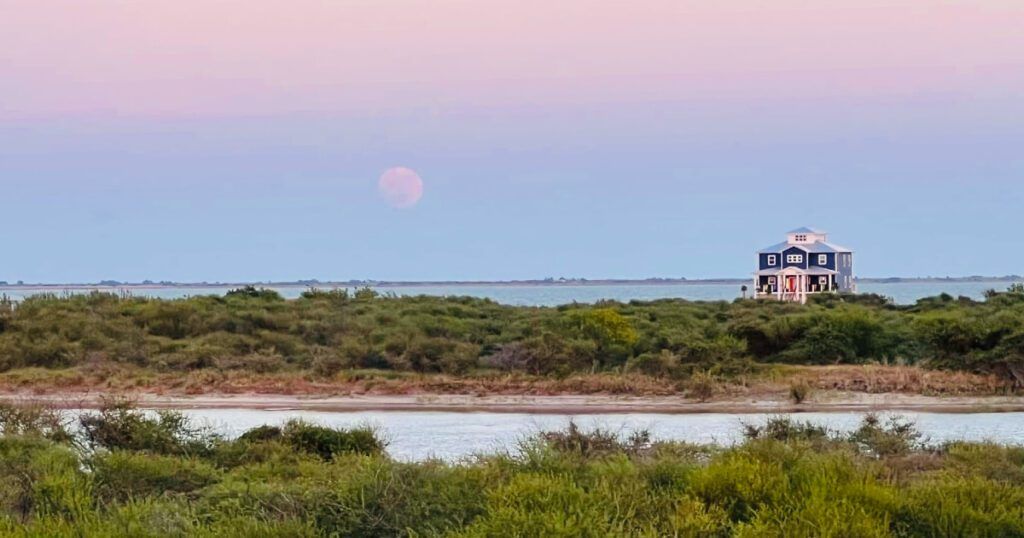 A picturesque scene of a blue coastal house, elevated on stilts, surrounded by lush greenery and overlooking calm waters at dusk. The sky is a soft gradient of pink and purple as the sun sets, with the moon faintly visible on the horizon, creating a serene and peaceful atmosphere.