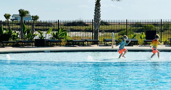 Two children wearing bright orange swim trunks play joyfully in a shallow pool under a sunny sky.