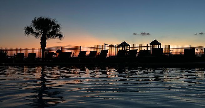 A tranquil sunset view by a pool, with the water gently reflecting the deepening hues of the sky. Silhouetted against the fading light are lounge chairs, a palm tree, and a fenced area, evoking a relaxing atmosphere. The warm orange and yellow glow on the horizon contrasts with the cool blue tones of the sky and water.