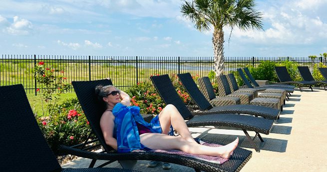 texas vs. florida-woman sitting by pool-3 A woman relaxes on a lounge chair by a pool, wrapped in a blue towel, enjoying a sunny day. She is wearing sunglasses and reclining with one hand resting on her face. The poolside area features several black wicker lounge chairs, and a neatly landscaped background with flowers, a palm tree, and a black metal fence. Beyond the fence, there is a grassy landscape under a bright blue sky dotted with fluffy clouds, adding to the warm and leisurely atmosphere.
