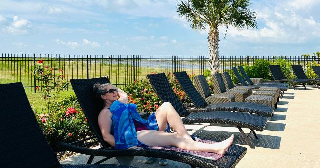 A woman relaxes on a lounge chair by a pool, wrapped in a blue towel, enjoying a sunny day. She is wearing sunglasses and reclining with one hand resting on her face. The poolside area features several black wicker lounge chairs, and a neatly landscaped background with flowers, a palm tree, and a black metal fence. Beyond the fence, there is a grassy landscape under a bright blue sky dotted with fluffy clouds, adding to the warm and leisurely atmosphere.