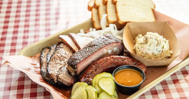 A tray of barbecued meats, including smoked brisket slices, ribs, and sausage, served with pickles, a cup of barbecue sauce, a small portion of potato salad, and slices of white bread. The tray is lined with butcher paper, and the meal is set on a red and white checkered tablecloth.