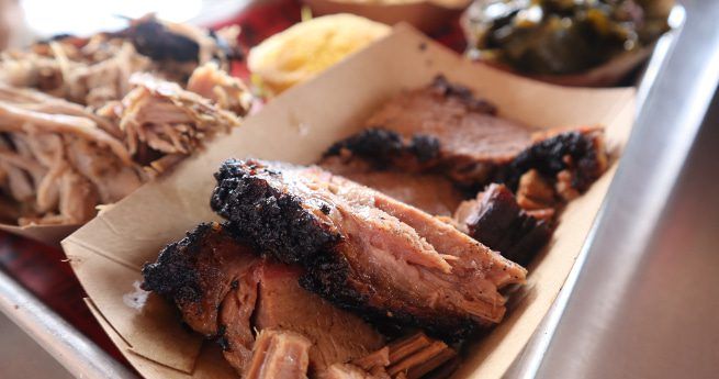 A close-up of tender slices of smoked brisket with a charred bark, served on butcher paper in a tray. Behind the brisket are sides of pulled pork, cornbread, and collard greens, creating a hearty barbecue meal. The tray is set on a metal surface, highlighting the rustic presentation of the food.