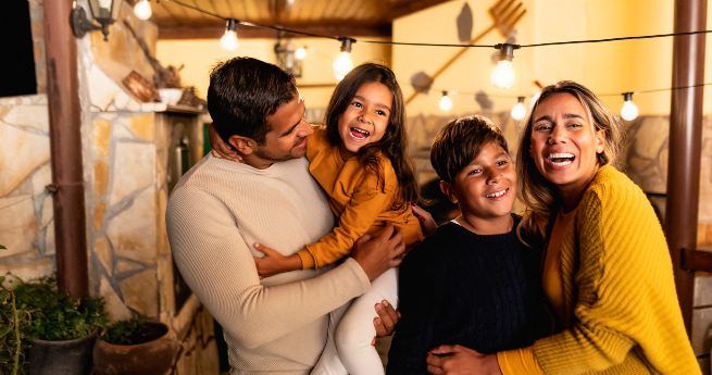 A cheerful family of four smiling and laughing while standing outdoors under string lights. The father is holding his daughter, and the mother is standing beside their son.