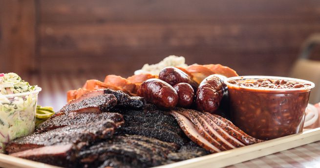 top bbq restaurants in victoria-large bbq plate-5 A large tray of assorted barbecued meats, including smoked brisket slices, ribs, sausage links, and sliced turkey, accompanied by sides of baked beans and coleslaw. The meal is served on a tray lined with butcher paper, with a red and white checkered tablecloth underneath, creating a classic barbecue presentation.