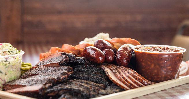 A large tray of assorted barbecued meats, including smoked brisket slices, ribs, sausage links, and sliced turkey, accompanied by sides of baked beans and coleslaw. The meal is served on a tray lined with butcher paper, with a red and white checkered tablecloth underneath, creating a classic barbecue presentation.