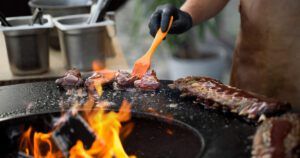 A person grilling ribs and meat over an open flame, using an orange basting brush to apply sauce. The grill is flat, with ribs and small pieces of meat cooking on it, surrounded by flames. The person is wearing a brown apron and black gloves, with metal containers in the background.