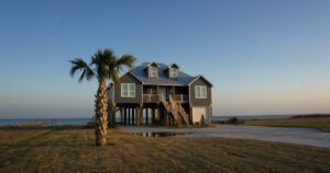 The image shows a raised coastal home with a gray exterior, standing on stilts, overlooking a calm beachfront.