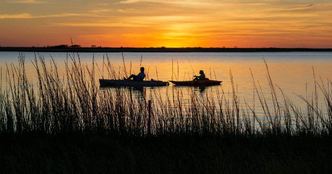 The image shows two people kayaking on a calm body of water during sunset. The golden and orange hues of the sky reflect off the water, creating a peaceful and serene atmosphere.