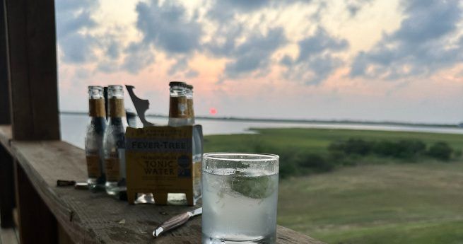 The image shows a refreshing drink in a glass placed on a wooden railing, with a scenic sunset view in the background. Several bottles of tonic water and a bottle opener are nearby, suggesting a relaxing moment outdoors.