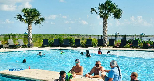 The image shows a group of people enjoying a sunny day at a swimming pool. Several adults and children are sitting or standing in the shallow end, socializing near the pool's edge. The pool area is surrounded by palm trees and a fence, with lounge chairs lined up in the background. Beyond the fence, lush greenery and a distant body of water create a scenic view.