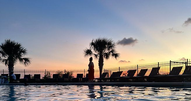 The image captures a serene poolside scene at sunset. A person stands near the edge of the pool, silhouetted against a colorful sky transitioning from warm orange to soft purple. Two palm trees frame the scene, with several lounge chairs lined up near a fence in the background.