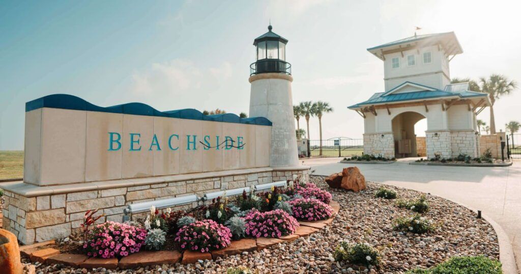 Entrance sign for a beachfront community named Beachside, featuring a lighthouse structure and a gatehouse in the background. The sign is made of stone and has the word 'BEACHSIDE' in blue lettering, surrounded by a well-maintained flower bed with vibrant blooms and decorative rocks.