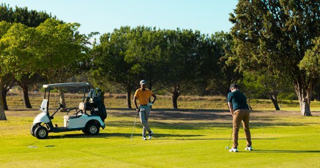 Two men playing golf on a sunny day, with one man putting while the other stands nearby holding a club. A golf cart is parked on the green, and trees line the background, creating a peaceful and scenic setting on the golf course.