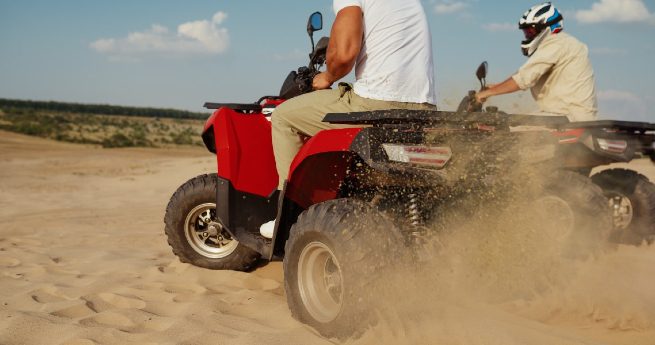 best places to live in texas-men on atvs-6 Two people riding all-terrain vehicles (ATVs) through a sandy landscape, kicking up dust as they move.
