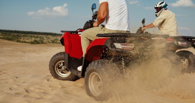 Two people riding all-terrain vehicles (ATVs) through a sandy landscape, kicking up dust as they move.