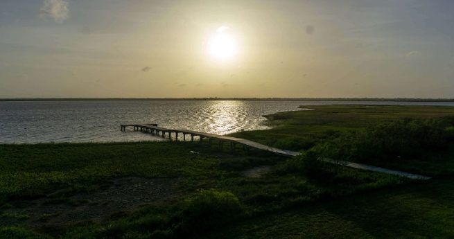 Serene sunset view over a calm body of water with a wooden pier extending into the water. The landscape features grassy marshlands and the soft glow of the setting sun reflecting on the water's surface, creating a peaceful and natural scene.