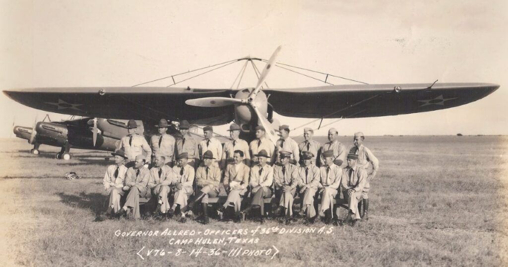 A vintage black-and-white photograph of a group of military officers posing in front of a single-engine aircraft at Camp Hulen, Texas. The plane features a large wing with visible propellers on either side, and the officers are dressed in formal military uniforms. They are seated and standing in two rows, with "Governor Allred, Officers 36th Division A.S. Camp Hulen, Texas" written at the bottom of the image, dated 8-14-36.