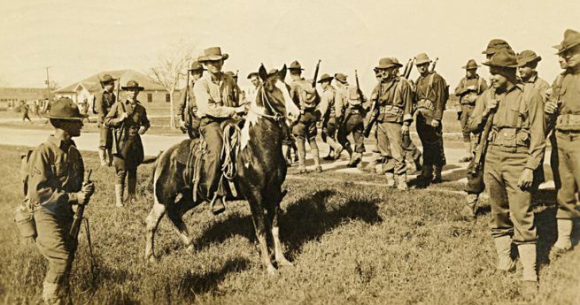 A sepia-toned photograph depicting a group of soldiers at Camp Hulen, Texas, standing in a loose formation. One soldier is mounted on a horse at the center of the image, while the others, dressed in military uniforms and helmets, are standing nearby, holding rifles.